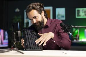 Tech guru promoting mechanical keyboard from sponsor to subscribers. Content creator paid by brand to showcase computer peripheral device with neon lit keys in home studio photo