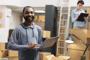 African american man overseeing product shipment and order logistics, working in small scale factory operations with taped boxes stacked on shelves. Handling goods and processing orders. photo