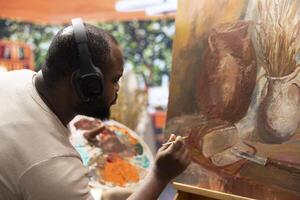 Inspired artist working on a canvas with music on headset, painting a rustic artwork using fine bristles to apply dye with precision. Creating a masterpiece with supplies in art and craft atelier. photo