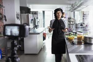 Skilled chef in apron recording a vlogging session on plating food, stands behind the kitchen counter. Expert offers explanations and details as she teaches techniques for the food industry. photo