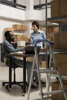 Diverse team at desk discussing order processing to guarantee distribution in a tightly packed storage room filled with products and boxes. Organized structure of a small scale business. photo