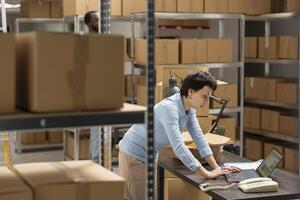 Young woman at her desk reviewing shipment data for delivery services, verifying taped boxes in a storage room before sending them for distribution. Hand packing goods for retail startup. photo