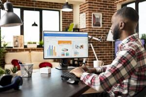 Remote male manager seated at home desk, organizing fiscal data and analyzing company progress report on computer. African american man formulates financial strategies for small business development. photo