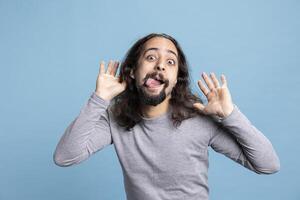 Playful young man with a long hair and beard making a goofy face in the studio. Carefree guy radiates humor and joy, capturing a lighthearted moment full of laughter and casual personality. photo