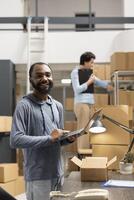 Warehouse employee using laptop to coordinate orders and ensure the distribution activity, small business logistics process. Preparing taped boxes across the organized supply room. photo
