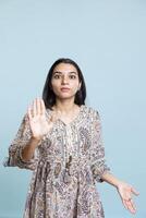 Focused firm indian woman showing stop hand symbol and complaining in studio. Authoritative person doing firm rejection symbol with her palm, having enough against blue background. photo