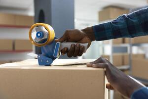 Factory worker applies adhesive tape to boxes while attaching air waybill and tracking numbers. African american staff manages packing, labeling and express delivery process in warehouse. photo