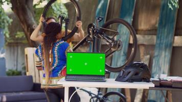Laptop showing greenscreen while healthy and active interracial pair fixes bicycle components. Minicomputer with isolated copyspace screen on table while couple does bike maintenance at home yard. photo