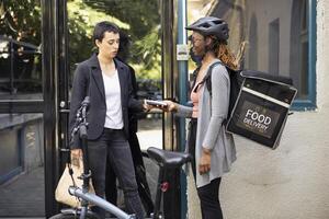 Woman completes a fast food purchase with contactless payment at the doorstep, courier holds pos terminal steady as the electronic transaction is processed. Electronic wireless purchase. photo