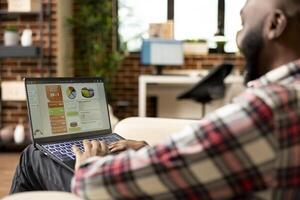 African american man using digital device to analyze marketing plans and draft business reports at home. Black male entrepreneur sitting on couch, focused on project presentation on laptop. photo
