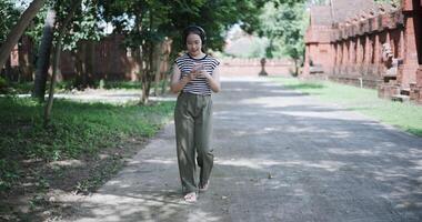 Handheld shot, Asian woman listening to music on wireless headphones over a mobile app while walking leisurely in a quiet place. video