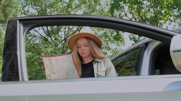Blonde woman in hat staying next to car door checks the route on map to get to the destination. Slow motion Young tourist explore local travel making candid real moments. True emotions expressions of getting away and refresh relax on open clean air video