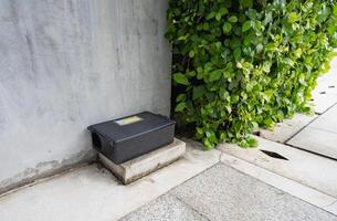 Tamper proof black rodent bait station placed on concrete ground near green foliage. Modern residential pest control for safe and effective urban rat infestation prevention. Secure rat bait box. photo