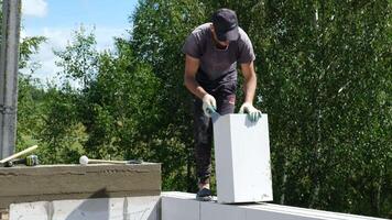 A young Caucasian man with short hair works on a construction site. He is applying mortar to a white block while standing on a wall. Green trees are in the background. video