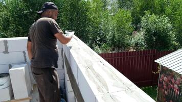 A man works on a construction project, applying mortar to a wall. He is wearing a cap and casual clothing. The background features greenery and a wooden fence. video