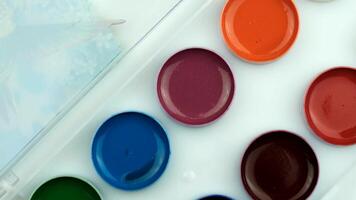 A close-up of a watercolor paint palette with various vibrant colors arranged in circular wells. The background is light and neutral, enhancing the colors. video