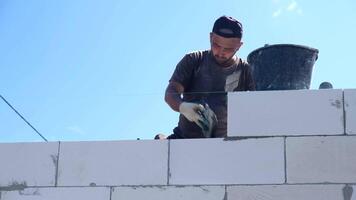 A young Hispanic man is working on a construction site, laying blocks for a wall. He wears a cap and gloves, with a bucket nearby. video