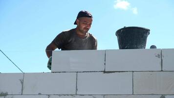 A young Hispanic man is building a wall with white blocks. He is wearing a cap and working with a bucket nearby. The sky is clear and blue. video