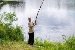 Boy standing on lakeside with a long stick, playing near the water in nature on a summer day photo