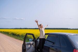 Happy boy standing with raised arms on country road in rapeseed field near car on sunny summer day, joyful outdoor adventure photo