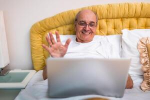 Senior man in pajamas sitting on bed, smiling and waving during call on his laptop, enjoying remote work from home. photo