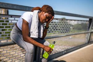Tired young sporty woman takes a break from running to rehydrate with energy drink while sitting on bridge with towel around her neck. photo