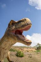 Huge Artificial dinosaur tyrannosaurus, close-up of dinosaur head against sky background. With copy space. Dinosaur has open mouth, you can see its teeth, front legs. Yellow rocks in the background photo