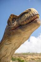 Huge Artificial dinosaur tyrannosaurus, close-up of dinosaur head against sky. With copy space. Dinosaur shows its teeth, yellow rocks and trees in the background photo