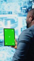 Vertical African american tech engineer reviews algorithmic data next to mockup, holding a tablet surrounded by digital visuals. AI smart technology and neural networks in a modern environment. Camera A. video