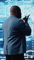 Vertical African american IT specialist calling a technician on his smartphone, talking about automation via neural networks systems. Businessman discuss about coding and data processing. Camera A. video