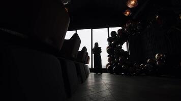 Silhouette of Woman Standing at Window During Evening Event, Black and Gold Balloon Arch in Luxurious Indoor Venue With Dim Lights and Reflective Floor in video