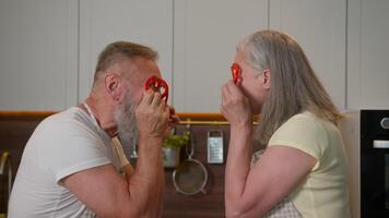 Funny happy retired grandma grandpa Caucasian old man woman having fun together make red paprika glasses peppers circles middle-aged wife husband old grandparents cooking fresh vegetables in kitchen video