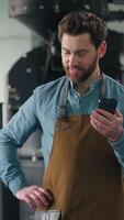 Vertical view Caucasian man working in roasted coffee fabric factory inspecting roasting machine caffeine beans checking quality holding mobile phone smartphone smiling happy technologist male job video