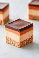 Square layered sweets on plain surface. Brown base, orange layer, light middle, dark top. Gluten free dessert. Low carb. Tasty treats. photo