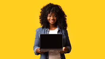 Cheerful beautiful black woman using laptop with empty screen over yellow studio background, copy space. Cute african american young lady showing notebook with blank display, mockup photo