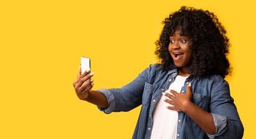 Emotional african american young woman with brand new mobile phone screaming with joy over yellow studio background, panorama with copy space. Amazed black lady holding modern smartphone photo