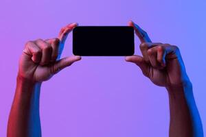 Black Male Hands Holding Smartphone With Black Screen In Horizontal Orientation Under Vivid Neon Light Over Purple Background, Mockup Image With Copy Space For Your App, Game Or Website Design photo