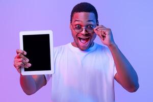 Check This App. Surprised African Guy In Sunglasses Holding Digital Tablet With Black Blank Screen For Mockup, Demonstrating Copy Space For Your Design, Standing In Neon Light On Purple Background photo