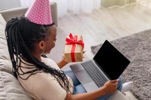 Young black woman with party hat and present using laptop with empty screen at home, copy space. African american lady having online party, showing gift box to blank computer screen, mockup photo