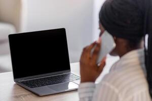 Unrecognizable black woman having conversation on mobile phone, using laptop with blank screen, copy space, back view. African american freelancer talking with clients on smartphone while using pc photo