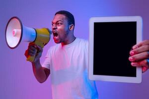 Emotional Black Guy Holding Digital Tablet With Blank Screen And Shouting To Loudspeaker, Making Announcement About New Website Or App, Standing In Vivid Neon Light Over Purple Background, Mockup photo