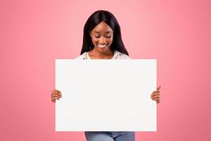 Awesome promo. Happy African American woman holding empty banner with empty space on pink background. Beautiful black lady showing blank poster, mockup for your design photo