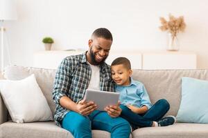 Black Dad And Son Using Digital Tablet Browsing Internet And Watching Cartoons Online Spending Time Together At Home. Weekend With Daddy. Family Gadgets Concept photo
