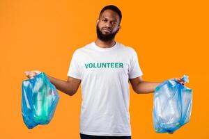 Displeased Black Male Volunteer Holds Two Plastic Bags Full Of Litter And Garbage Standing Over Yellow Background. Studio Shot. Ecology Problems, Waste Sorting Concept photo