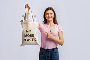 Eco-Activism Concept. Young Smiling Woman Pointing At Tote Bag With No More Plastic Inscription, Collecting Empty Bottles For Reuse And Recycling, Standing Over Grey Studio Background, Copy Space photo