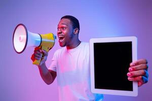 Excited African Guy Making Announcement With Loudspeaker And Holding Digital Tablet With Black Blank Screen, Standing Over Purple Background Under Neon Lighting, Mockup Image With Copy Space photo
