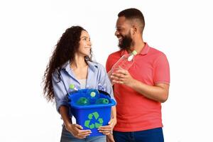 Waste Sorting. Multicultural Family Couple Sorting Junk Posing Holding Box With Recycle Symbol Full Of Used Plastic Bottles Standing Over White Background. Studio Shot. Trash Disposal Concept photo