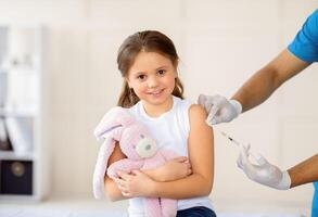 Happy little girl with toy bunny getting coronavirus vaccine injection at hospital. Black doctor immunizing child against covid-19 virus at clinic. Healthcare and medical concept photo