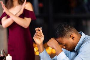 She Said No. Woman Rejecting Marriage Proposal From Her Black Boyfriend, Upset African American Man Sitting At Table In Restaurant With Diamond Ring In Hand, Touching Head In Despair, Selective Focus photo