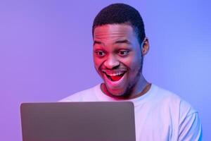 Closeup Shot Of Excited Black Guy Using Laptop Computer Under Neon Lighting, Joyful African American Man Looking At Computer Screen With Amazement, Standing Over Purple Background, Free Space photo